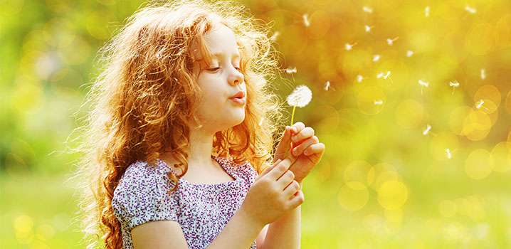 Mädchen mit lockigen Haaren steht im Sonnenlicht und pustet auf eine Pusteblume