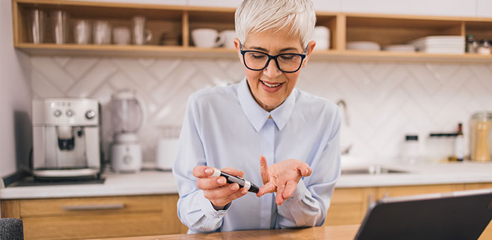 Ältere Frau sitzt in ihrer Küche und testet sich mit einem Messgerät auf Diabetes