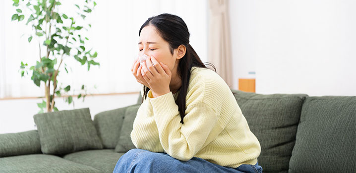 Junge Frau sitzt vornübergebeugt auf der Couch und hält sich ein Taschentuch an die Nase