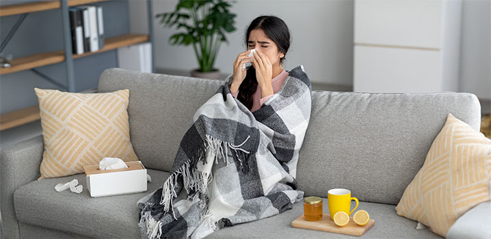 Junge Frau sitzt in Decke gewickelt auf dem Sofa und schnäuzt sich die Nase