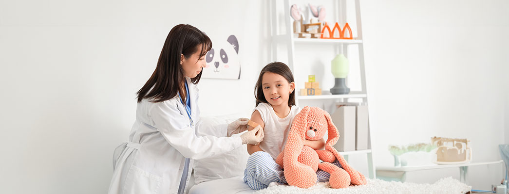 Junges Mädchen sitzt mit einem Kuscheltier auf dem Bett und bekommt ein Pflaster