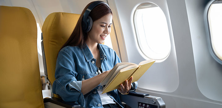 Junge Frau sitzt am Fenster im Flugzeug, trägt Kopfhörer und liest ein Buch