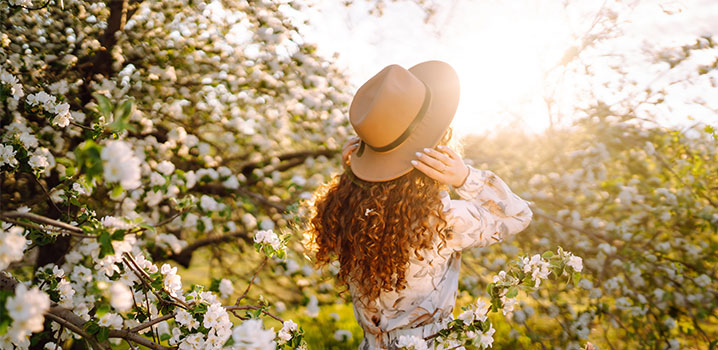 Junge Frau mit lockigem Haar und Strohhut steht mit dem Rücken zur Kamera zwischen blühenden Bäumen