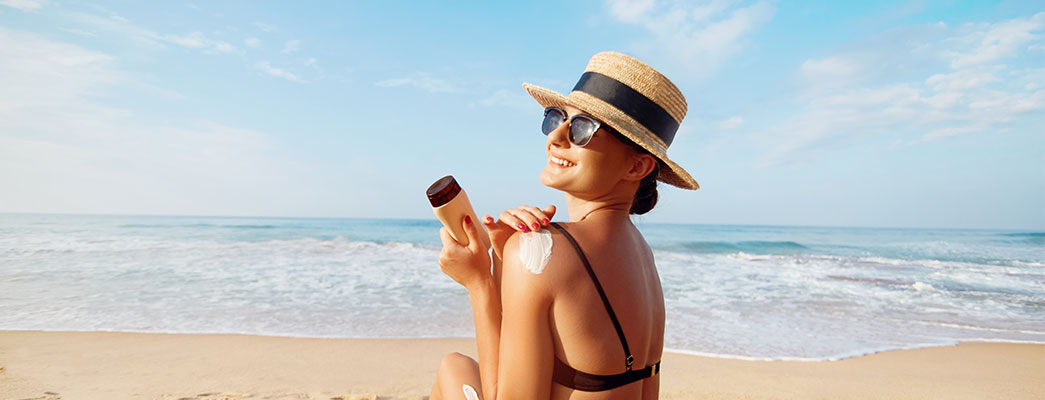 Junge Frau im Bikini und mit Sonnenhut sitzt am Strand und cremt sich die Schulter ein