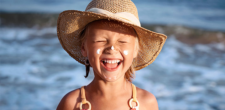 Mädchen mit Strohhut am Strand, kneift die Augen zusammen und lächelt
