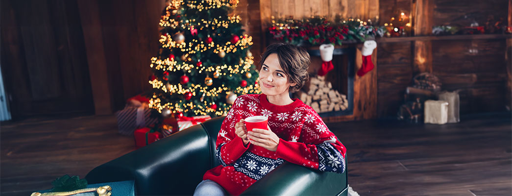 Frau im Weihnachtspulli sitzt mit einer Tasse in der Hand vor dem Weihnachtsbaum